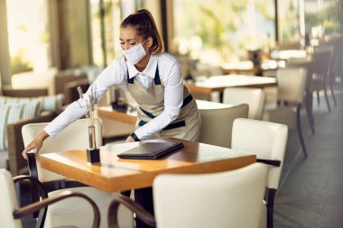 Waitress wearing protective face mask and adjusting chairs and tables in a cafe. 
