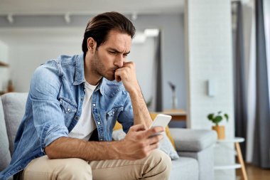 Young worried man sitting on the sofa and reading text message on mobile phone. 