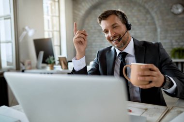 Happy businessman drinking coffee while making video call over laptop in the office.