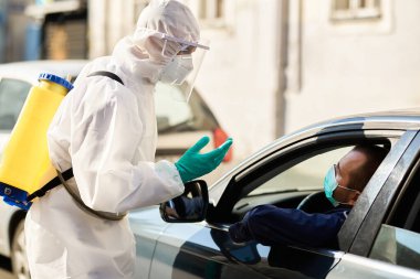 Sanitation worker in hazmat suit communicating  with a man in car at drive through checkpoint during coronavirus epidemic. 