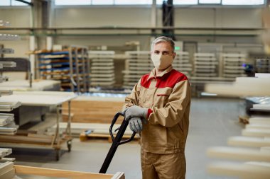 Portrait of a carpenter with face mask working with pallet jack in a warehouse and looking at camera. 
