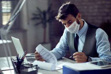 Male entrepreneur analyzing business reports while wearing face mask and working in the office during virus epidemic. 