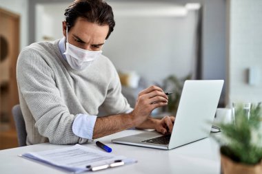 Young businessman with protective face mask using laptop while working on  reports at home. 