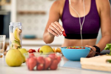 Close-up of athletic woman adding strawberries while making fruit salad in the kitchen. 