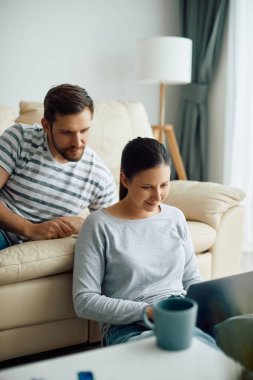 Smiling woman using laptop while relaxing with her husband at home. 