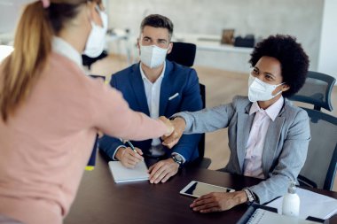 Members of human resource team wearing protective face masks while having job interview with potential candidate in the office.  Focus is on black businesswoman fist bumping with a candidate. 