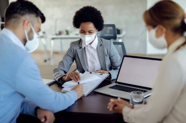 African American bank manager pointing at place of signature on a contract while closing a deal with a couple during the meeting in the office. All of them are wearing protective mace masks due to COVID-19 epidemic. 