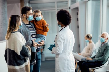 Young parents with small daughter communicating with their family doctor at medical clinic during coronavirus pandemic. 