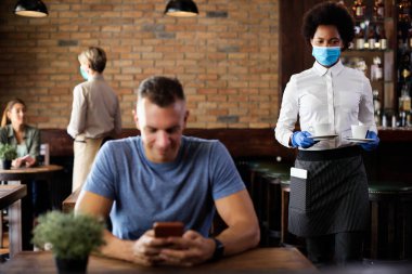African American waitress serving coffee and wearing protective face mask and gloves due to COVID-19 pandemic.