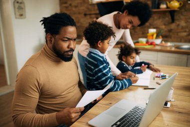 African American man using computer and working at home while his wife is homeschooling their children. 