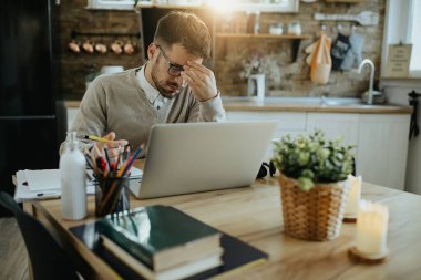 Male entrepreneur holding his head in pain while working on laptop at home. 