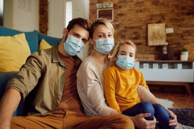 Portrait of parents and their daughter wearing face masks while sitting at home during COVID-19 pandemic. 