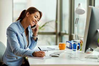 Young happy businesswoman talking on mobile phone and taking notes in the office. 
