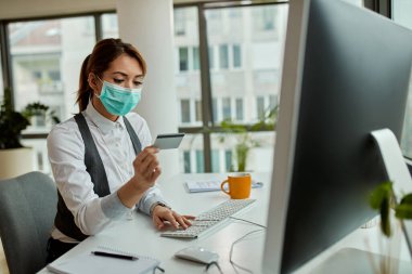 Businesswoman with face mask using credit card while checking her online bank account on a computer in the office. 