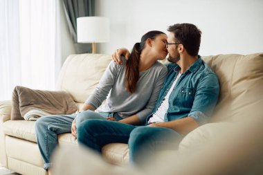 Couple in love embracing and kissing while sitting on the sofa in the living room. 