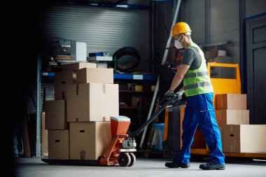 Warehouse worker with face mask pushing packages on pallet jack at storage compartment.