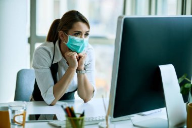 Young businesswoman wearing face mask while using desktop PC and reading problematic e-mail in the office.