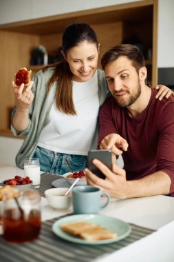 Young happy couple looking at something on mobile phone while eating breakfast together at home. 