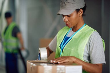 African American warehouse worker using tape dispenser while preparing boxes for shipment in a storage room.