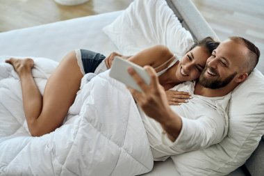 High angle view of happy couple taking selfie with smart phone while relaxing on a bed. 