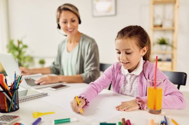 Smiling little girl enjoying and drawing on the paper at home while her mother is working on laptop.