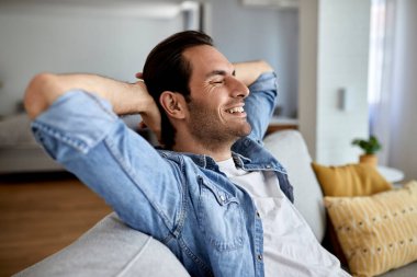 Young happy man with hands behind head relaxing on the sofa at home. 