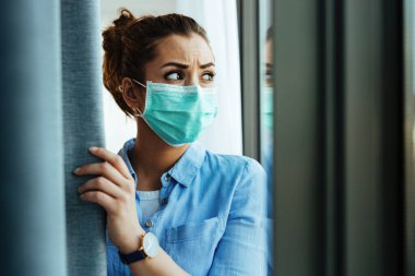 Distraught woman with protective mask on her face looking through the widow at home.