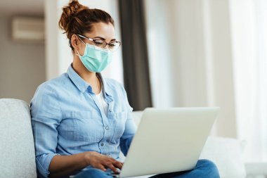 Young smiling woman with face mask using computer while relaxing in the living room.