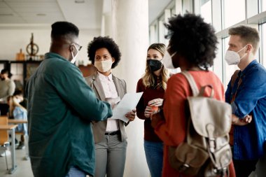 African American professor communicating with her students at university hallway. They are wearing face masks due to coronavirus pandemic.