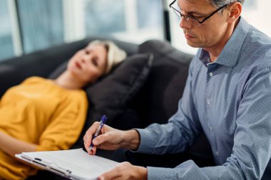 Male psychiatrist writing notes while having appointment with female patient at his office. clipart