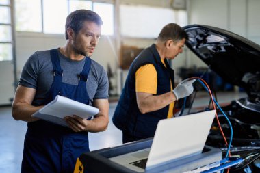 Two mechanic examining vehicle breakdown in auto repair shop. Focus is on mechanic using laptop while writing notes on clipboard. 