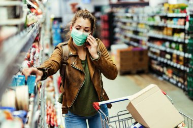 Young woman wearing protective face mask while buying groceries and talking on the phone during coronavirus pandemic in the supermarket. 