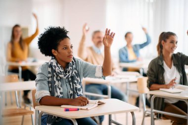 African American mid adult woman and her classmates raising their hands to answer a question during a class at lecture hall.