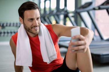 Happy athlete using cell phone and reading text message while taking a break from exercising at health club. 