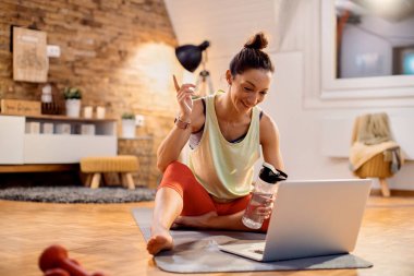 Happy sportswoman talking to someone via video call over laptop while sitting on the floor at home. 