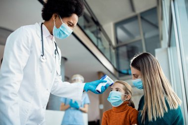 African American female doctor measuring temperature of a little girl who came to medical appointment with her mother during coronavirus pandemic.