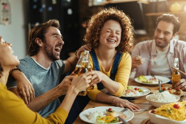 Group of young happy people laughing and having fun while toasting with beer during lunch at dining table. 
