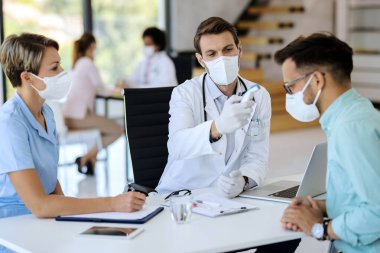 Male doctor wearing face mask while using infrared thermometer and measuring temperature of young man at medical clinic. 