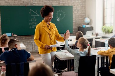 Happy African American teacher giving high-five to elementary student while congratulating her on good test results in the classroom. 