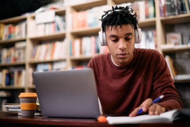 African American student using laptop and reading lecture notes while preparing for an exam at university library.