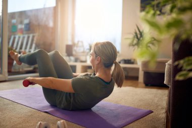 Female athlete using dumbbells while exercising sit-ups during home workout. 
