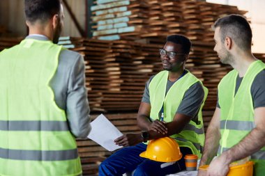 Happy warehouse workers communicating with quality control inspector at lumber department. Focus is on African American worker. 