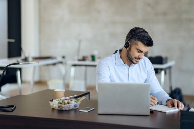 Young businessman with headset using computer and writing notes while working in the office. 