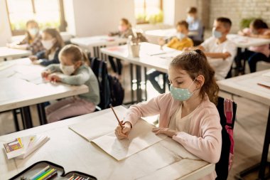 Schoolgirl wearing protective face mask and writing while studying with her classmates in the classroom. 
