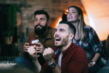 Young man and his friends cheering while watching rugby match on Tv at home.