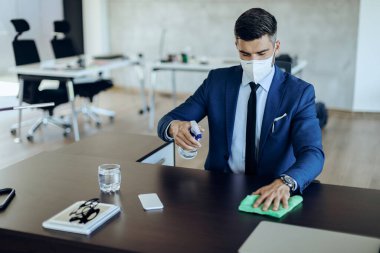 Businessman spraying his desk with disinfectant and wearing protective face mask while working in the office. 
