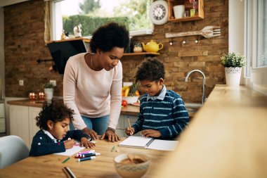 Black mother spending time with her kids who are coloring on the paper at home. 