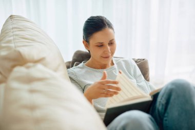 Smiling woman enjoying while reading a book and relaxing on the sofa in the living room. 