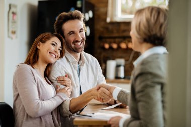 Young happy couple came to an agreement with their insurance agent. Focus is on man shaking hands with the agent. 