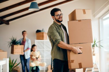 Happy father carrying stack of cardboard boxes while relocating in a new apartment. His wife and daughter are in the background. 